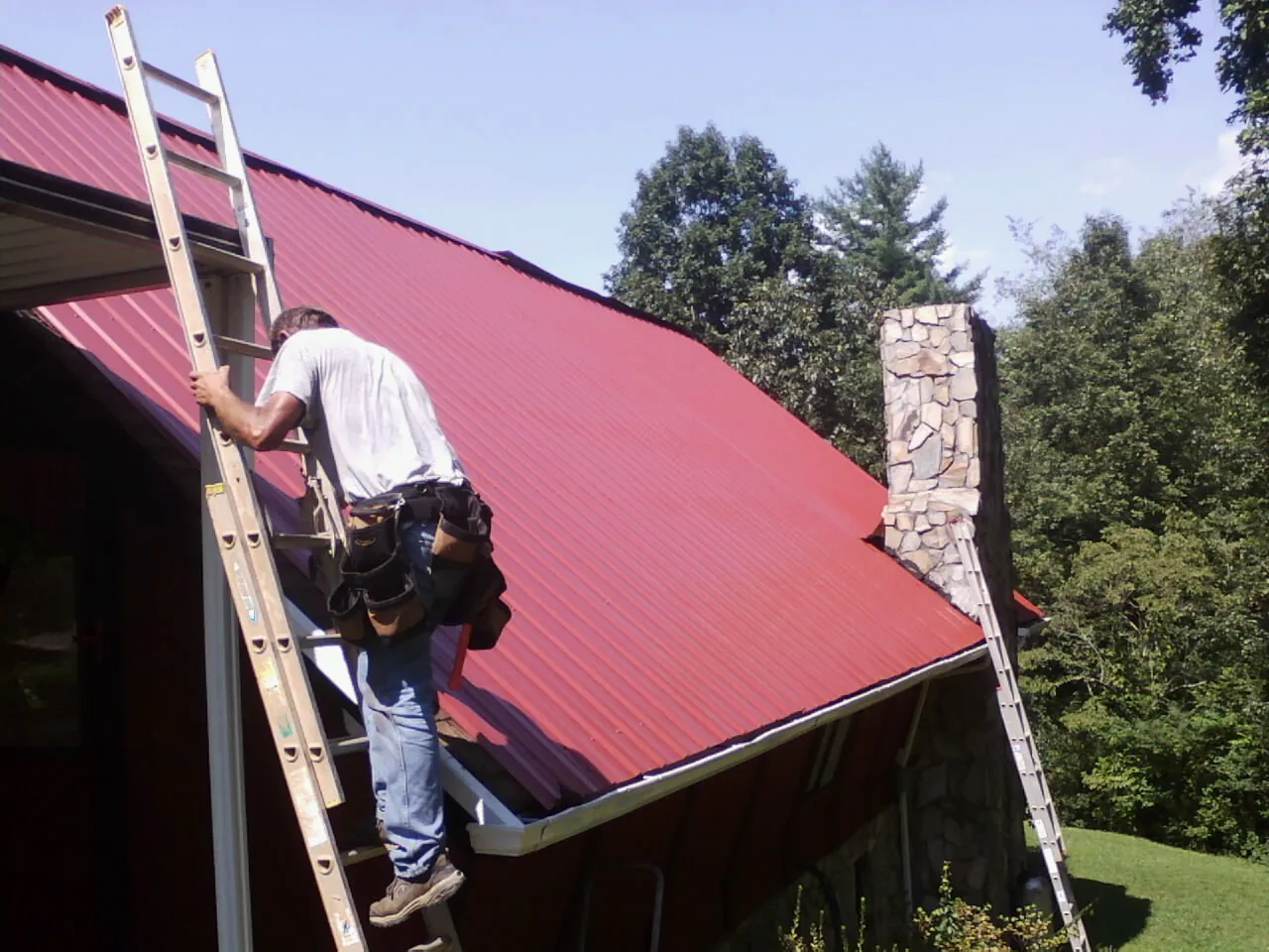 Roofer on a ladder beside a red metal roof and stone chimney — Soffit & Fascia Repair by Roofing Brothers in Milan